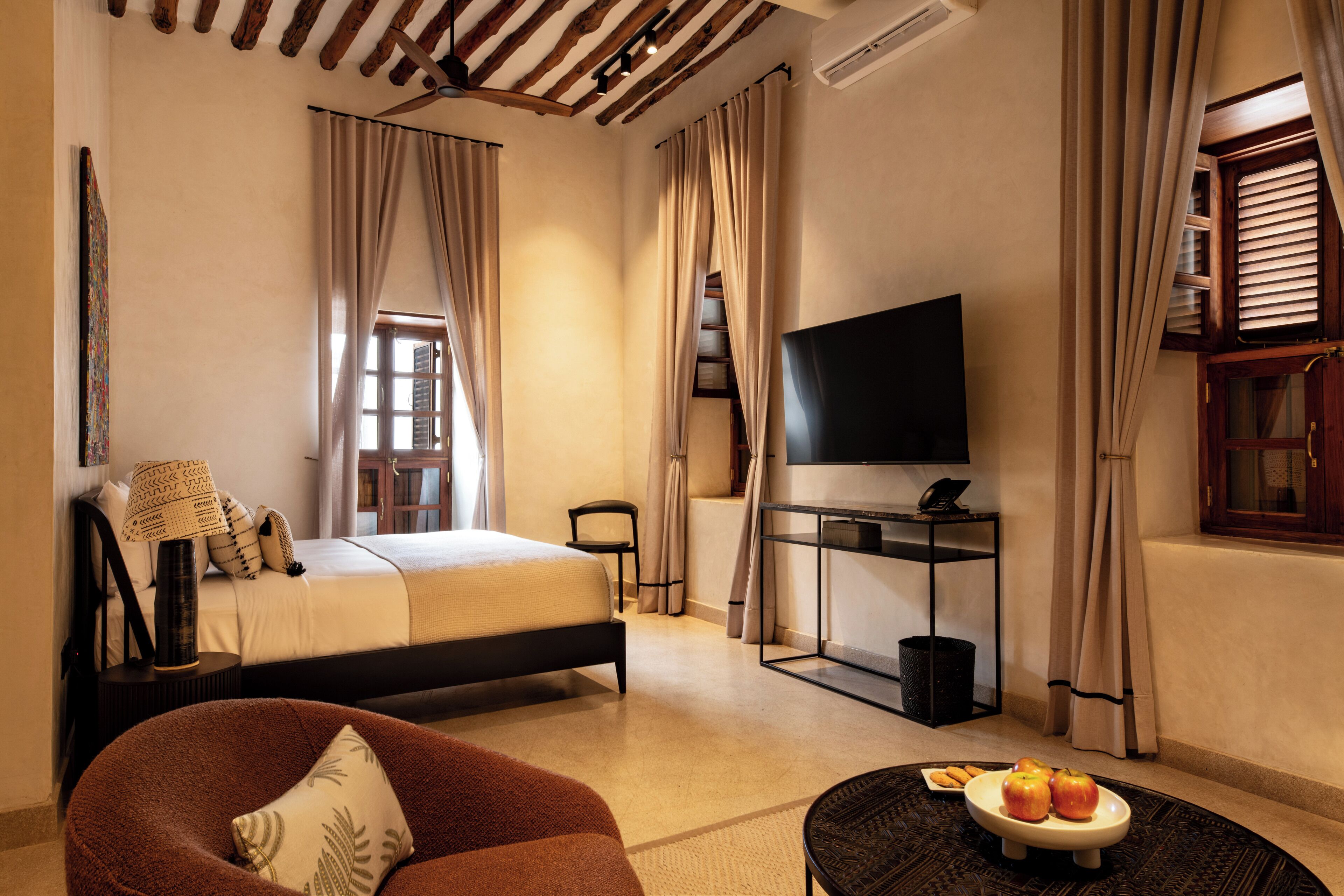A hotel room with a bed, a TV, a desk, a chair, a sofa, and a small table with fruits.