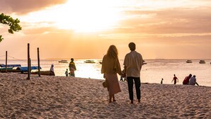 A beach scene with a couple walking hand in hand, boats in the water, and people in the background.