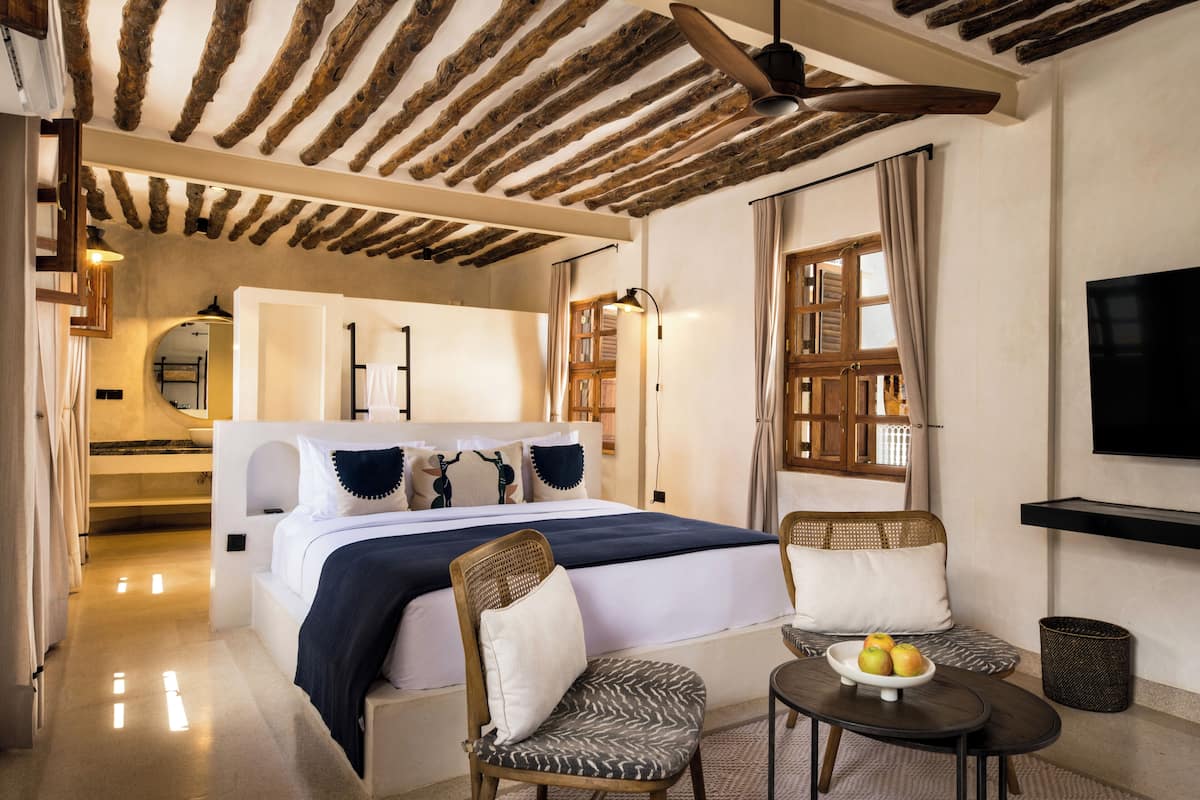 Habitación de hotel con cama, dos sillas, una mesita con fruta, televisión y techo con vigas a la vista.