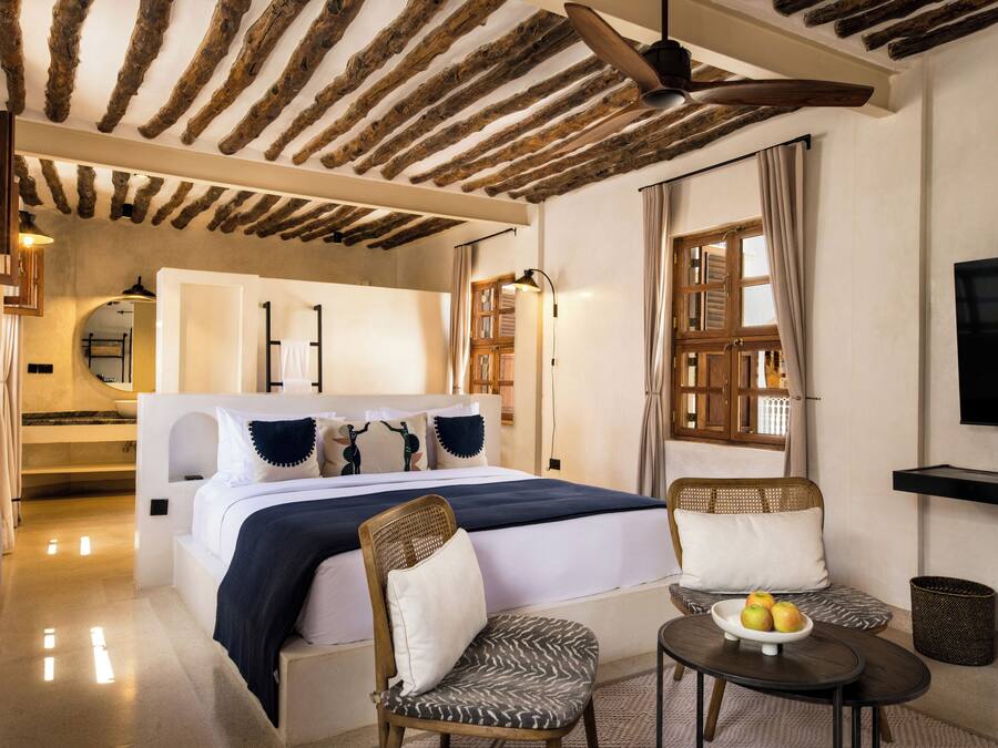 A hotel room with a bed, two chairs, a small table with fruit, a TV, and a ceiling with exposed beams.
