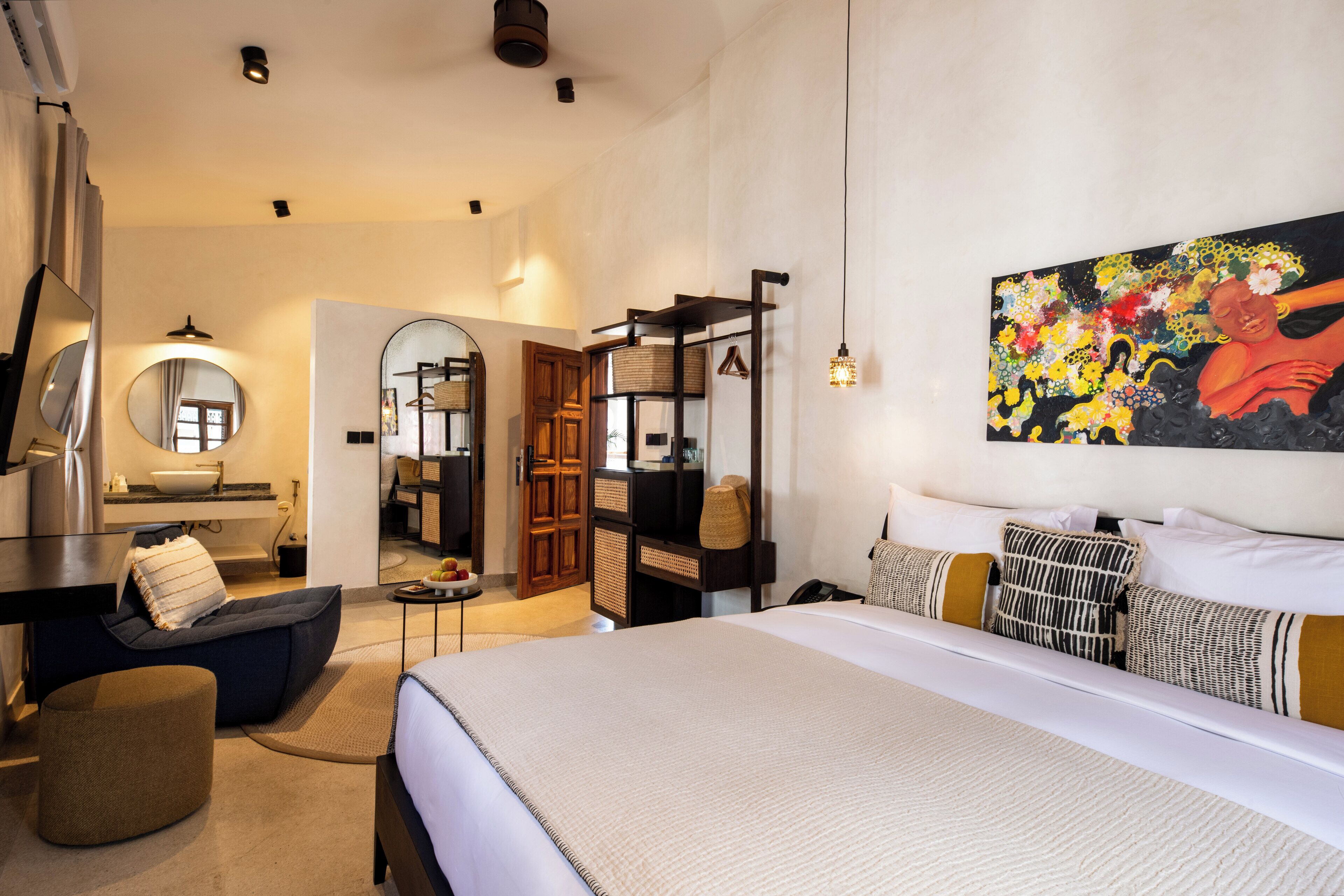 A hotel room with a large bed, a desk with a chair, a mirror, a wall-mounted shelf, and a painting.