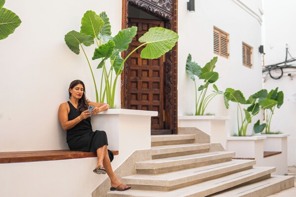 A person sitting on a bench outside a building with large green plants, a wooden door, and a staircase. - The Neela Boutique Hotel Stone Town (Zanzibar Town)