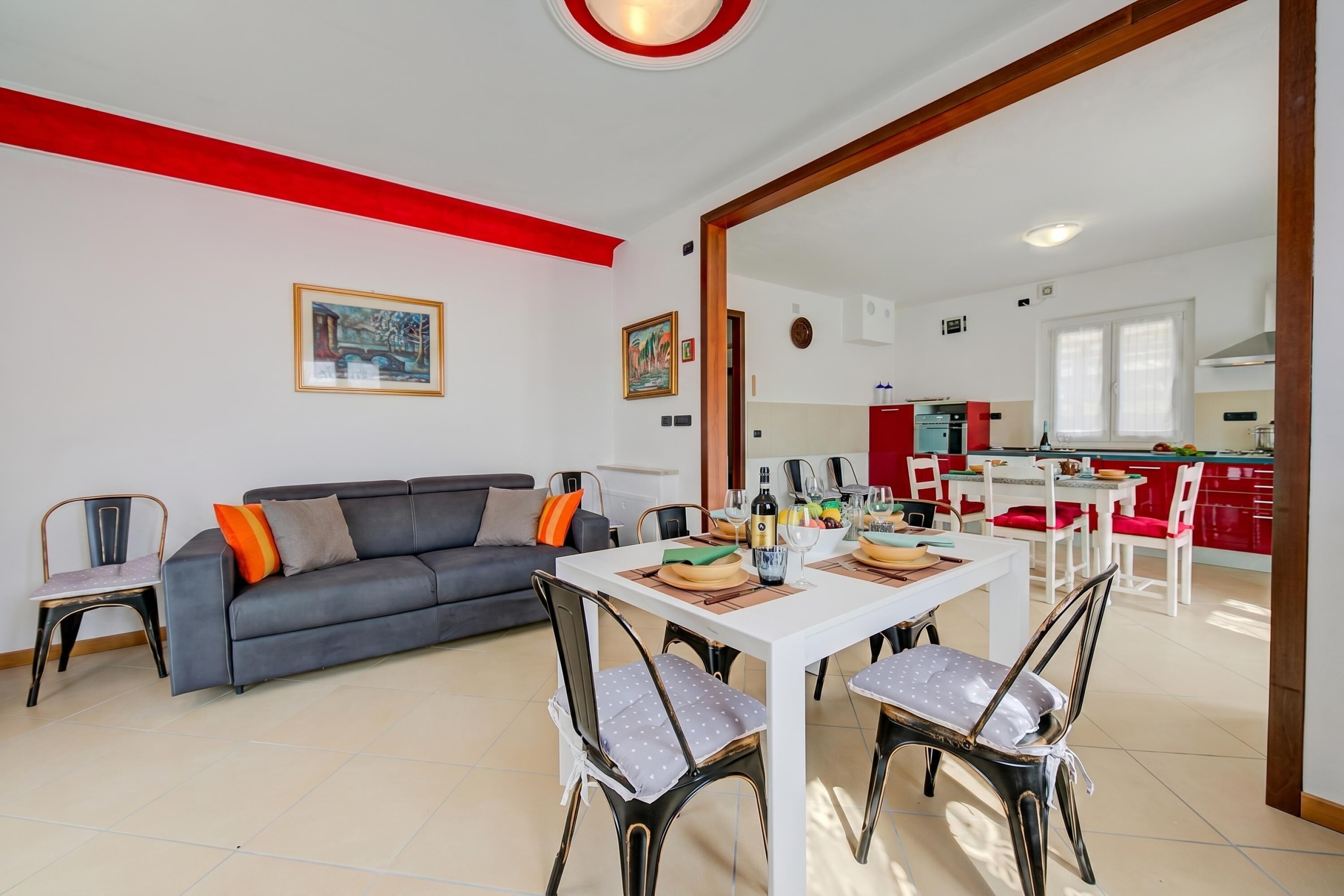 Un soggiorno moderno con un divano grigio, un tavolo da pranzo e sedie, una cucina sullo sfondo e una parete rossa a contrasto.