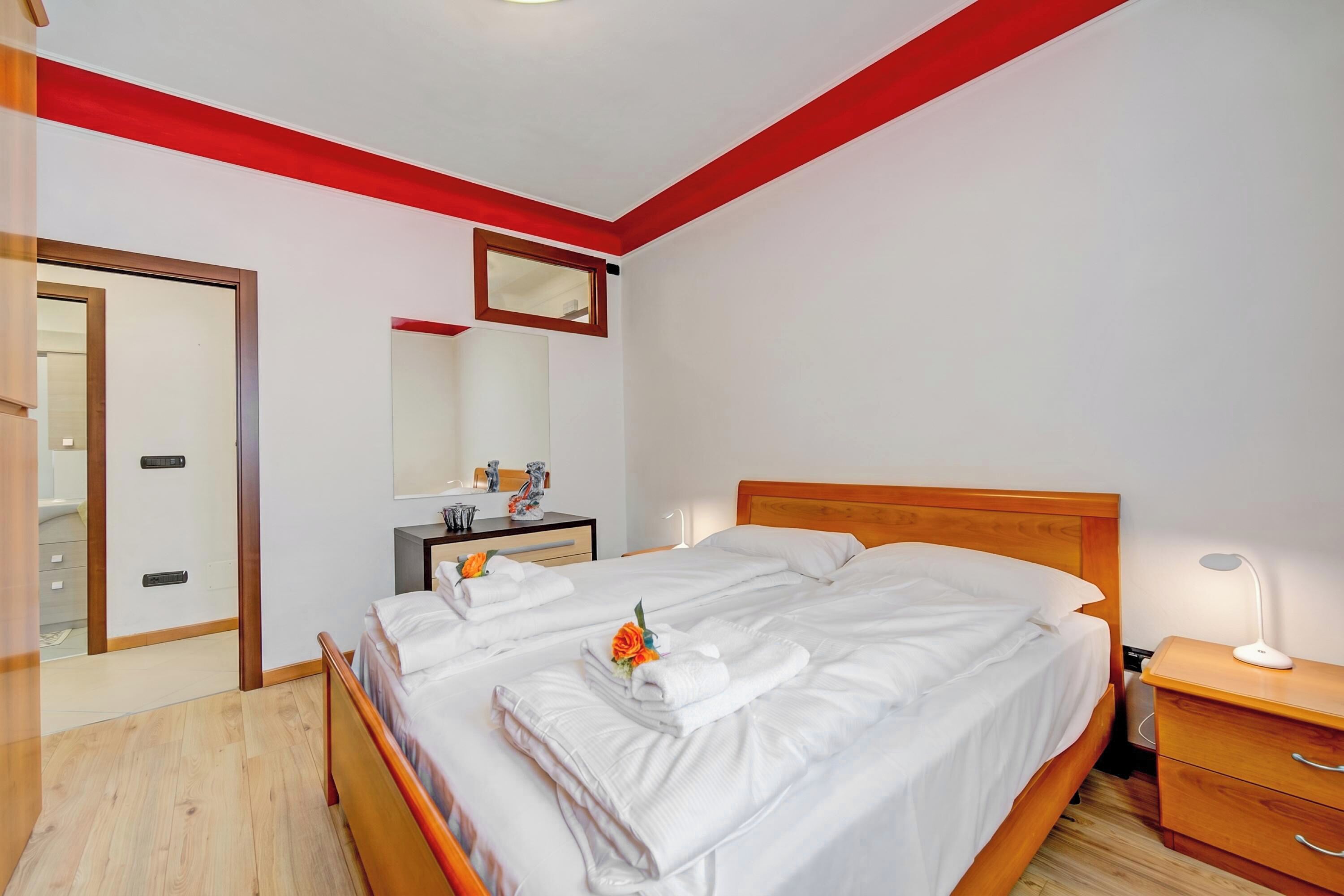 A hotel room with a wooden bed, a nightstand with a lamp, a mirror, and a door leading to another room.