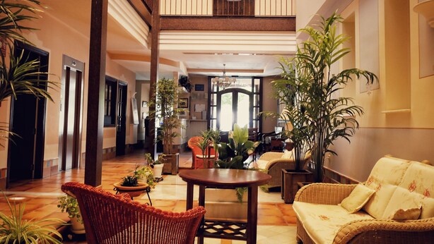 A spacious lobby with high ceilings, a central seating area with wicker furniture, and multiple potted plants. - Hotel Veracruz (Utrera)