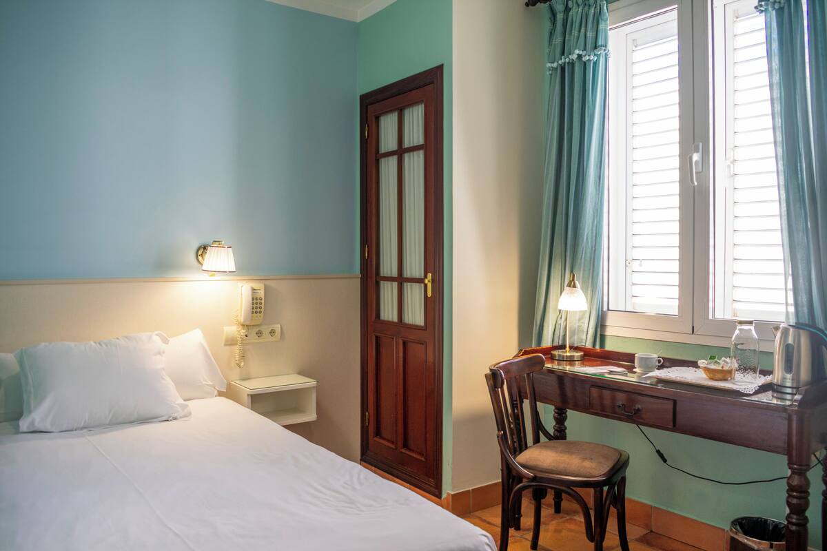 A hotel room with a bed, bedside lamp, desk with a chair, and a window with curtains.
