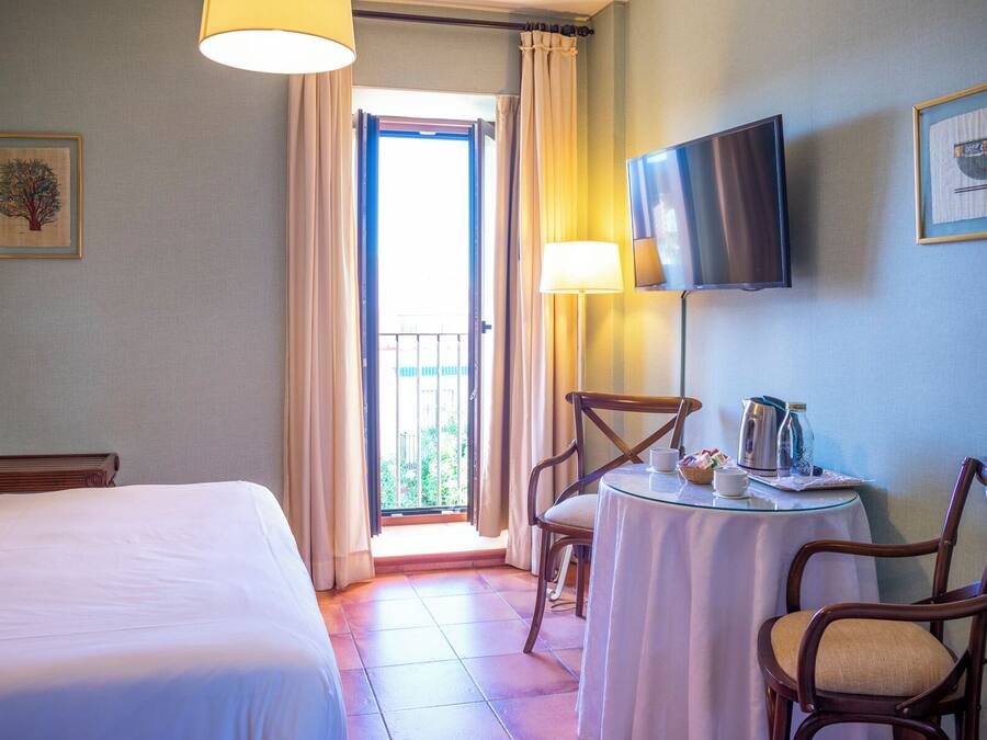 A hotel room with a bed, a table with chairs, a TV mounted on the wall, and a balcony with curtains.