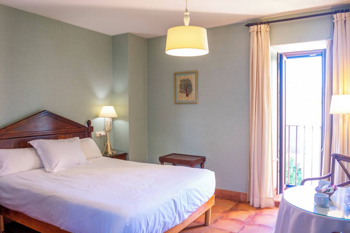 A neatly made bed with white linens, a wooden headboard, a bedside lamp, and a small table with a cup and tissues.