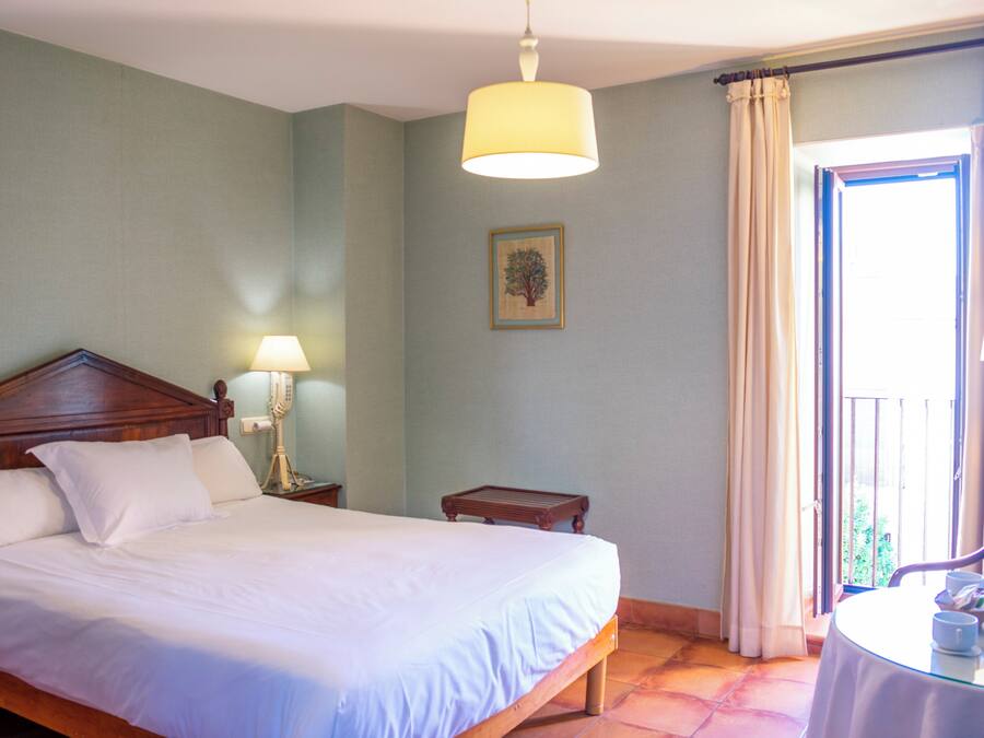 Uma cama bem arrumada com lençóis brancos, uma cabeceira de madeira, um abajur e uma mesinha com uma xícara e lenços.