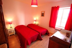 Una habitación con dos camas individuales, un aparador de madera, un escritorio con espejo y una ventana con cortinas rojas.