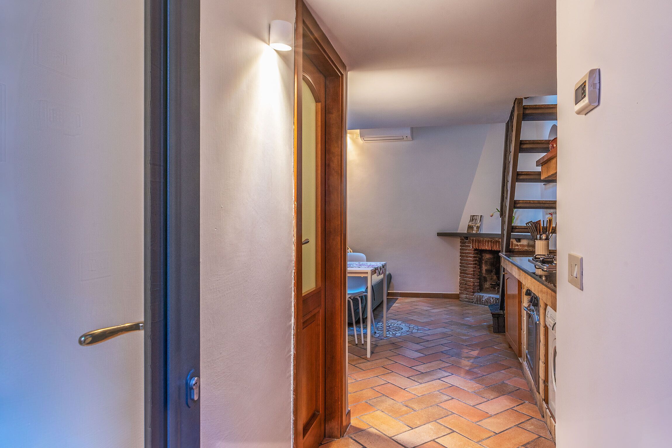 A narrow hallway with a wooden door, a small table with chairs, a tiled floor, and a kitchen area with a brick oven.