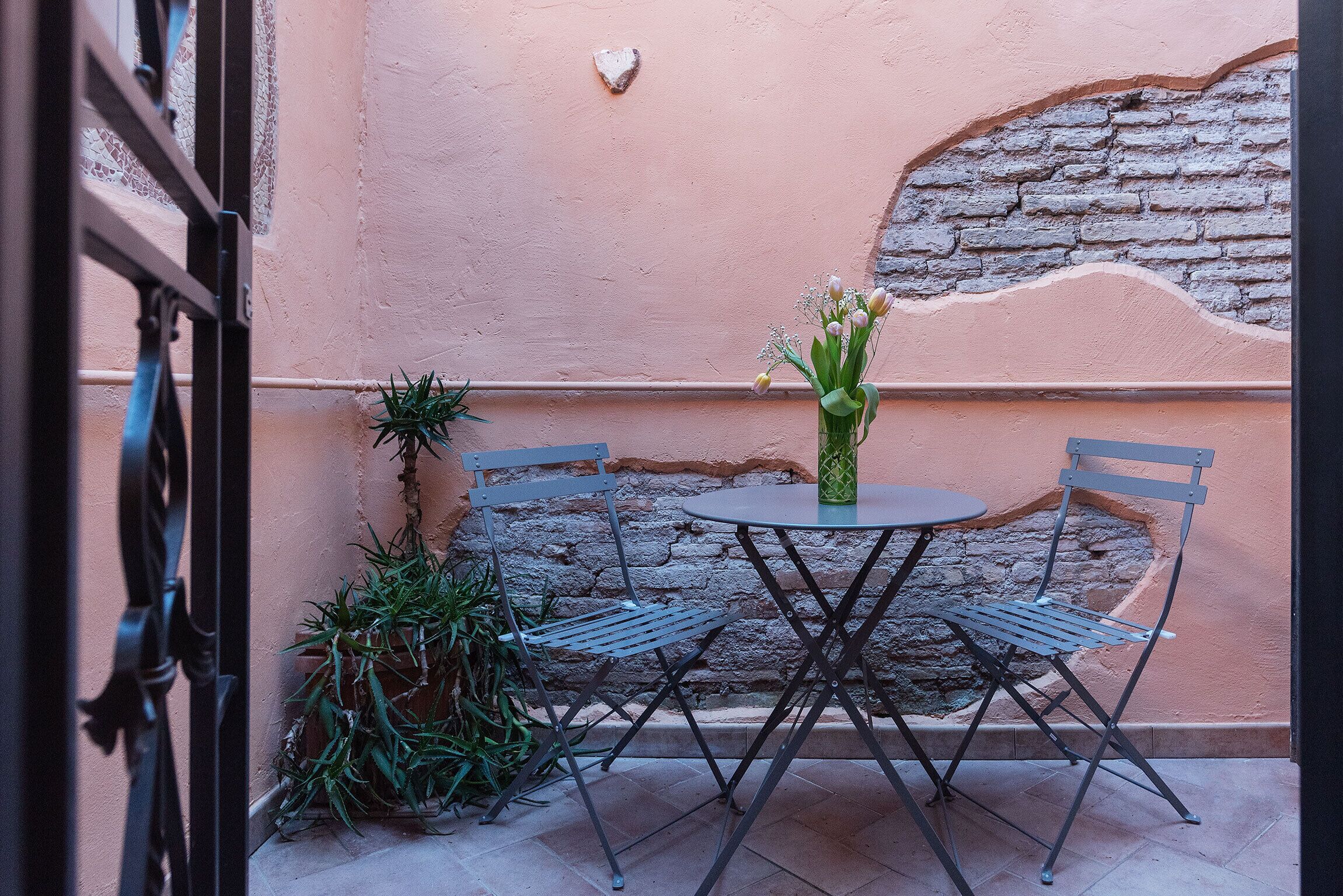 Une petite terrasse extérieure avec une table ronde et deux chaises, entourée de plantes en pot et d’un mur de briques.