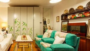 Una sala de estar con un sofá verde, una mesa de centro de madera y un armario blanco. Hay un espejo en la pared y una estantería con platos decorativos.