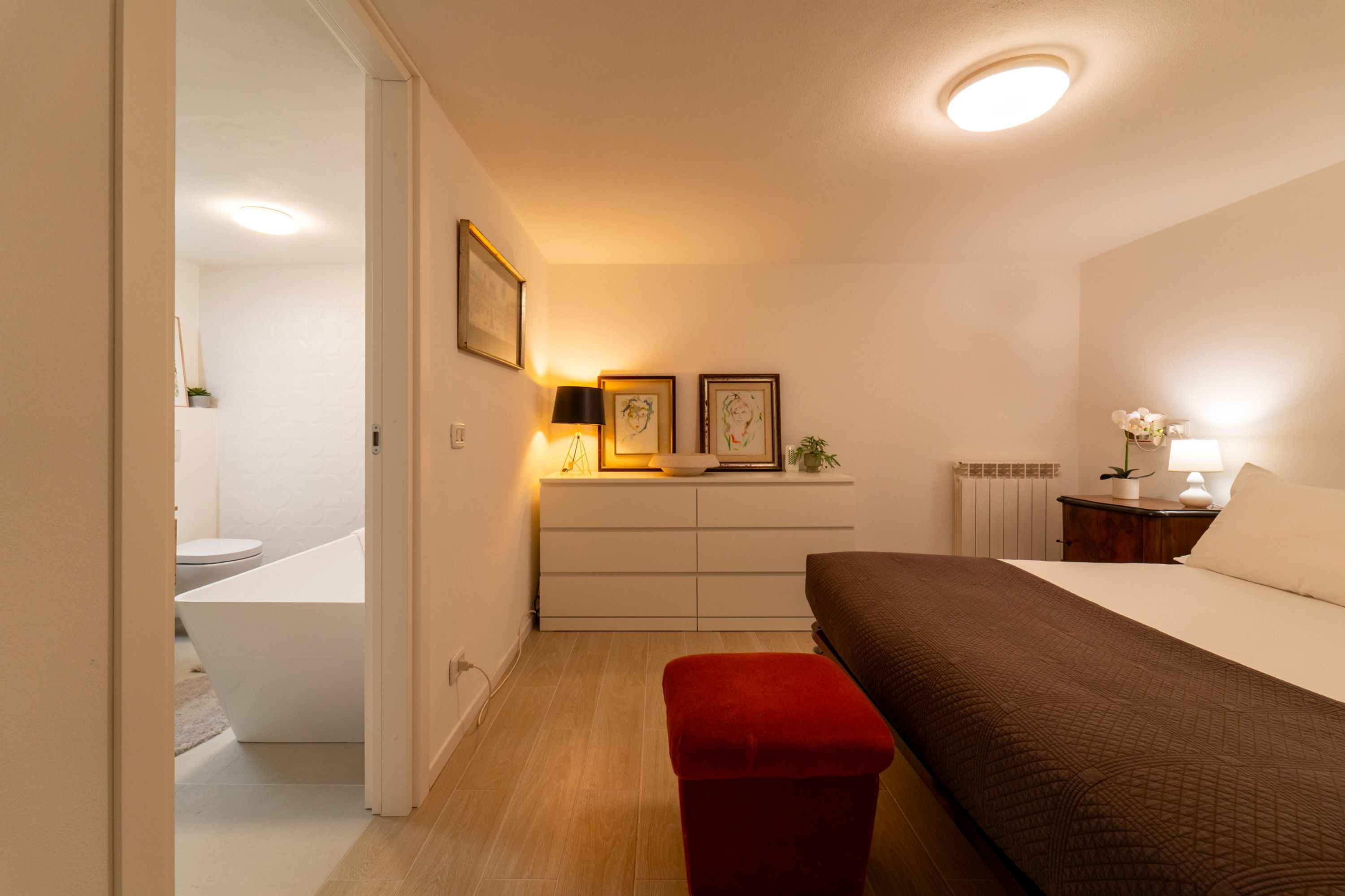 Une chambre à coucher avec un lit, un banc rouge, une table de chevet avec une lampe, une commode et des tableaux encadrés au mur.