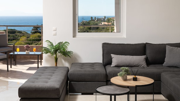 A modern living room with a grey sectional sofa, a round wooden coffee table, and a view of the sea. - The cozy nook (Chorafakia)