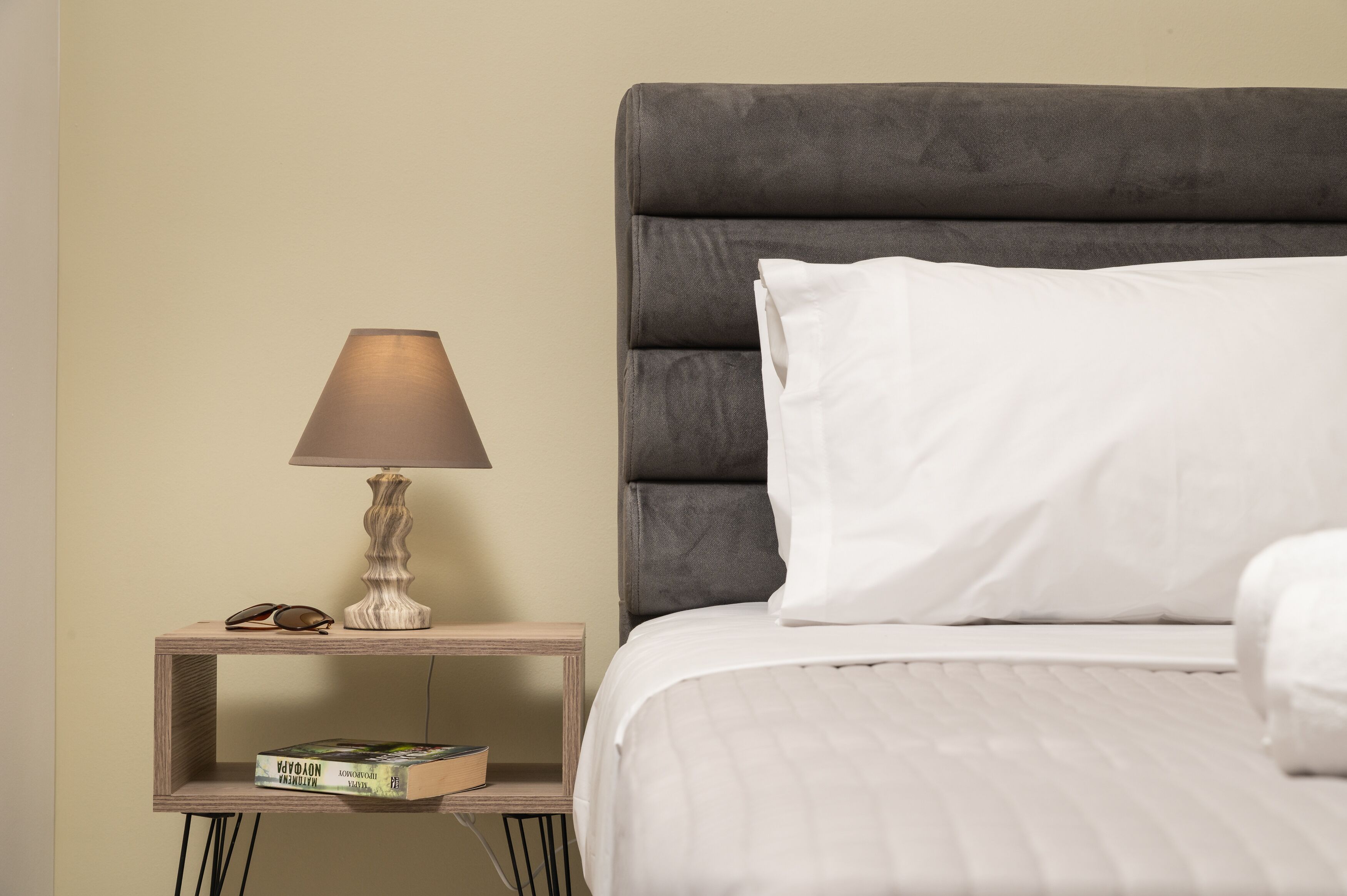 Una cama bien tendida con cabecera gris y almohadas blancas, una mesita de noche con lámpara y un libro en la estantería.