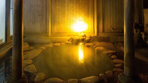 A hot spring bath surrounded by rocks, with a wooden wall and a light source in the background.