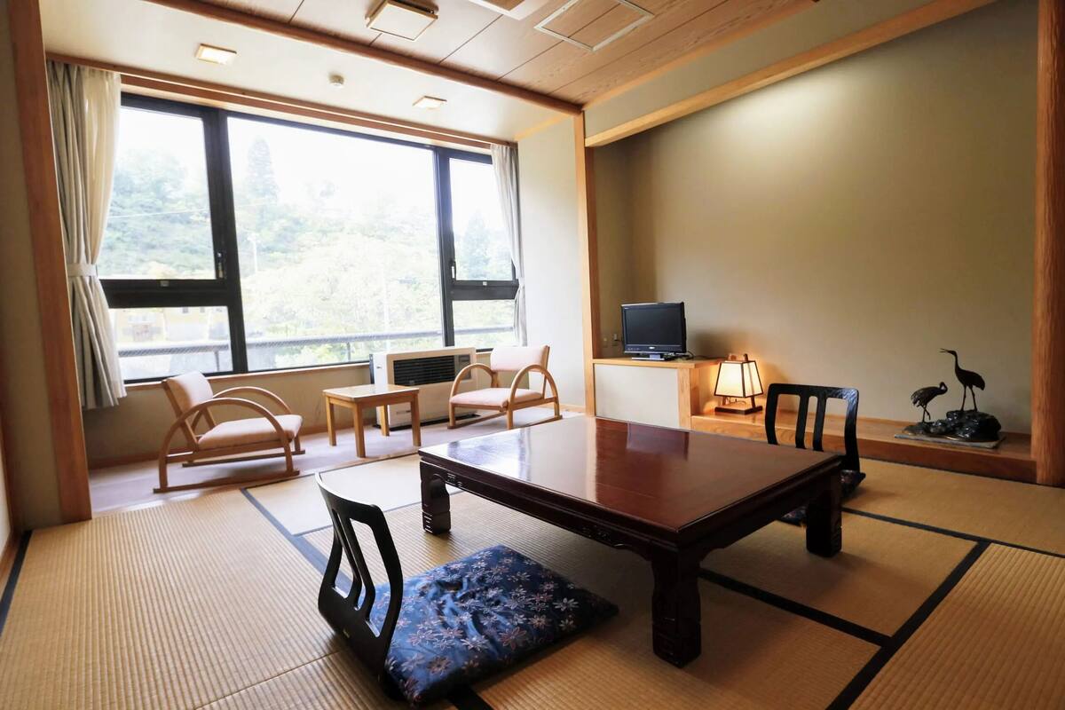 Une pièce de style japonais traditionnel, avec un sol recouvert de tatamis, une table basse, deux fauteuils rembourrés, un téléviseur et une statue d’oiseau décorative.