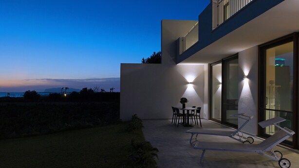 Un patio exterior moderno con una mesa y sillas de comedor, un sillón de descanso y un ventanal grande con vistas a un cuerpo de agua.