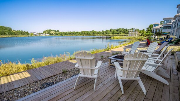 Ett trädäck med Adirondack-stolar med utsikt över en lugn sjö, omgivet av grönska och bostadshus.
