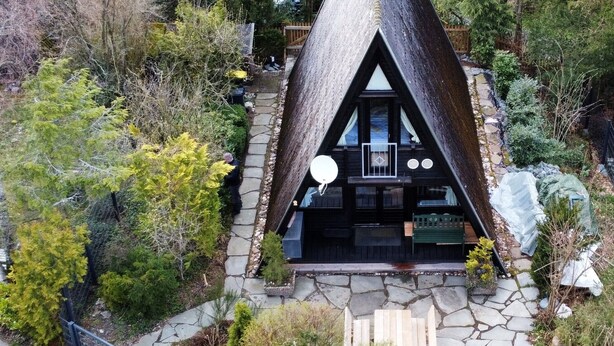 Une cabane au toit triangulaire, accessible par un sentier de pierres menant à l’entrée, entourée d’arbres denses et d’une végétation luxuriante.