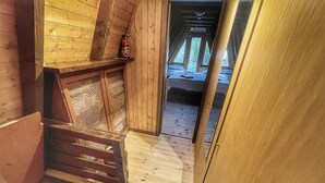 Un intérieur en bois avec un plafond en pente, un extincteur posé sur une étagère, un panier tressé et un lit donnant sur l’extérieur.