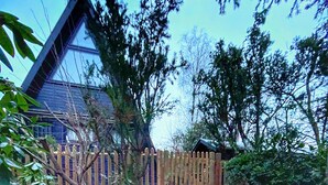 Une maison avec une clôture en bois, un escalier en pierre menant à la porte d’entrée et un jardin où poussent différentes espèces de plantes.