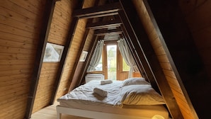 Une chambre confortable située sous les combles, avec un plafond en pente en bois, un lit recouvert de draps blancs, une table de chevet avec une lampe et une fenêtre agrémentée de rideaux.