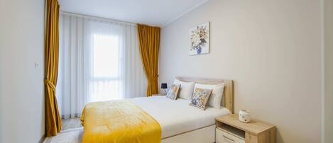 Una habitación de hotel con una cama grande, una mesita de noche con lámpara, un pequeño atril y una ventana con cortinas.