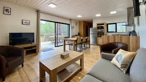 A modern living room with a flat-screen TV, wooden furniture, a sofa with a patterned cushion, and a dining area with a table and chairs. - New Ecogîte in the countryside, three minutes from all amenities. (Limoux)