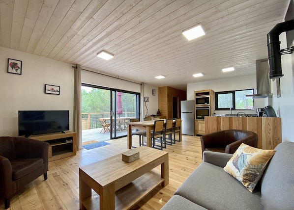 A modern living room with a flat-screen TV, wooden furniture, a sofa with a patterned cushion, and a dining area with a table and chairs.