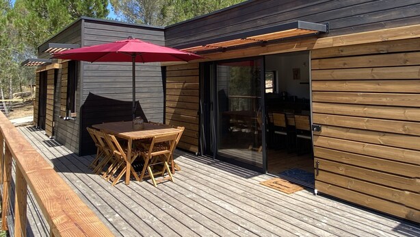 A wooden deck with a table and chairs, a red umbrella, and a sliding glass door leading to an interior space. - New Ecogîte in the countryside, three minutes from all amenities. (Limoux)