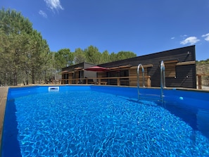 A swimming pool with a wooden deck, a red umbrella, and a modern building with large windows and wooden railings.