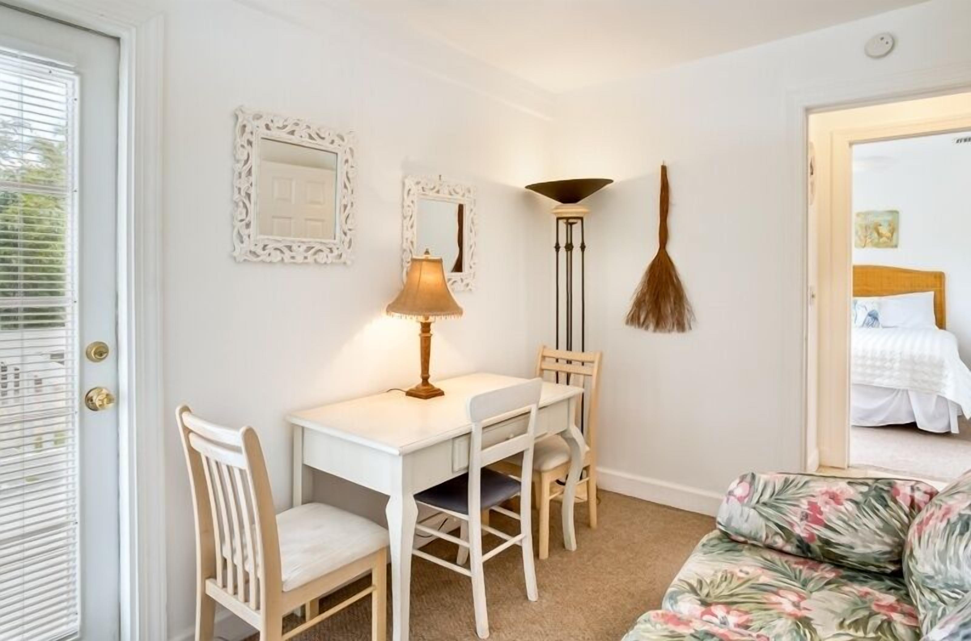 Ein Zimmer mit weißem Schreibtisch, zwei Stühlen, einer Lampe, einem Besen und einem Bett mit blumigem Bezug.