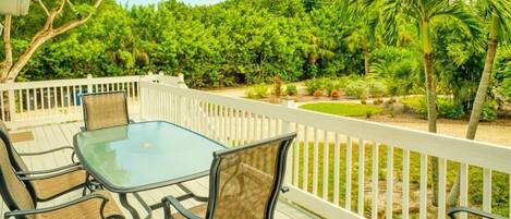 A deck with a glass table and chairs, surrounded by a white railing, overlooking a lush garden with various plants and trees.