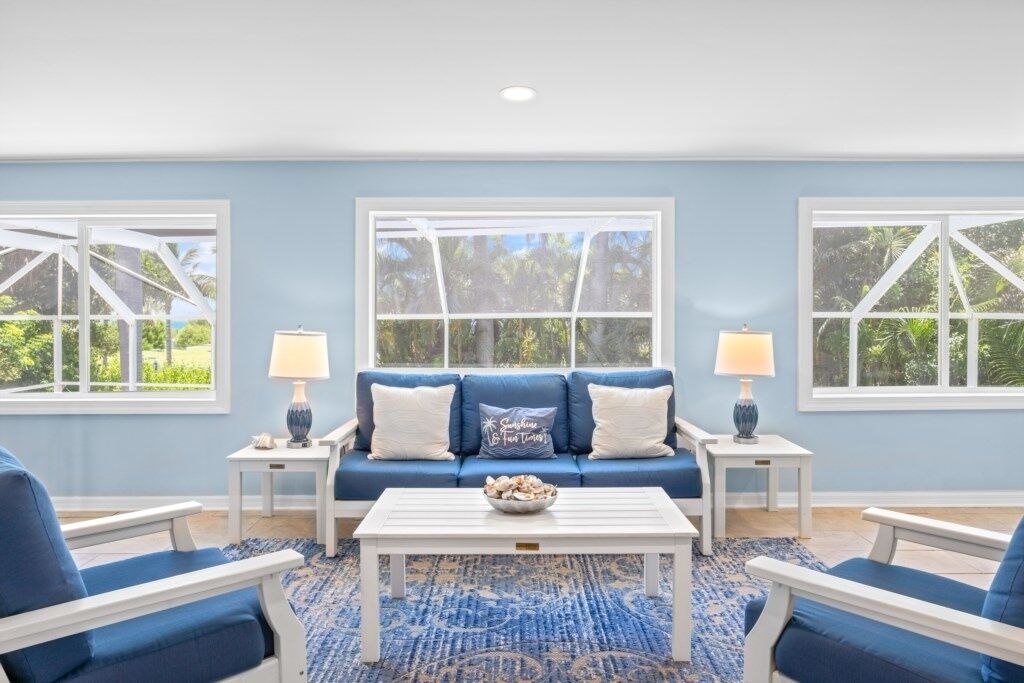 Quarto com um sofá azul, mesa de centro branca e cadeiras combinando. Há duas janelas com molduras brancas e vista para o verde lá fora.