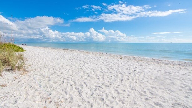 Ein sandiger Strand mit klarem, blauem Wasser und vereinzelten Wolken am Himmel.