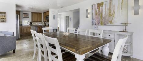 Un coin repas avec une table en bois et des chaises blanches, une cuisine en arrière-plan et un grand tableau abstrait accroché au mur.