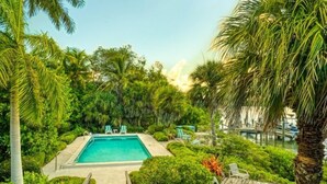 A rectangular swimming pool surrounded by lush greenery and palm trees, with a wooden deck leading to the pool and a dock with boats in the background. - Little Current 3 Bedroom Home by RedAwning (Captiva)