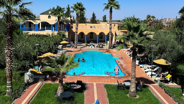 A swimming pool surrounded by palm trees and a building with arches. - Hotel Riad Qodwa (Sidi Abdallah Ghiat)