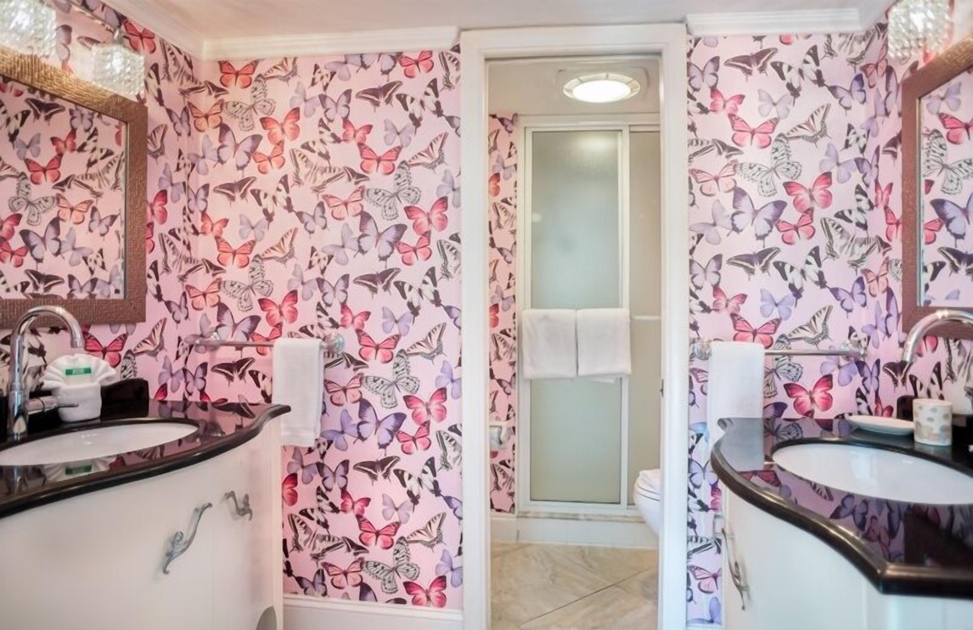 Un baño con papel tapiz estampado con motivos de mariposas, dos lavamanos y una puerta de ducha de vidrio.