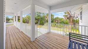 A covered porch with wooden flooring and white railings, overlooking a landscaped garden with palm trees and a clear sky. - Osprey House 4 Bedroom Home by RedAwning (Sanibel)
