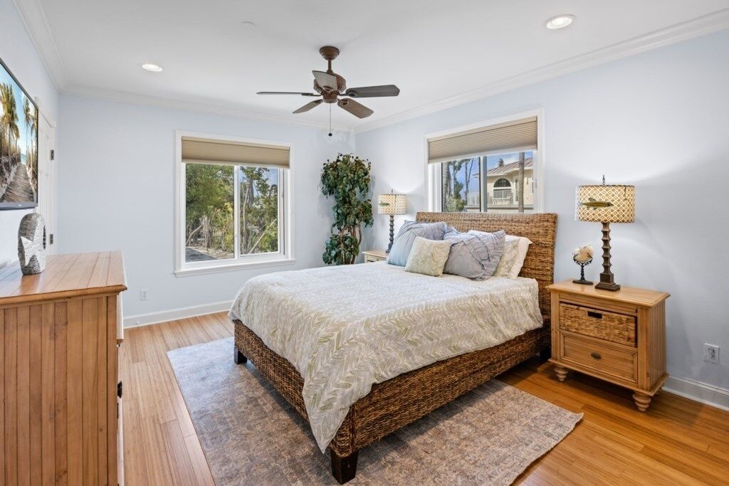 A bedroom with a large bed, wooden nightstands, a ceiling fan, and windows with views of greenery.