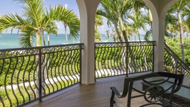 Een overdekt buitengebied met een houten terras, metalen hekwerk en uitzicht op de oceaan en palmbomen.