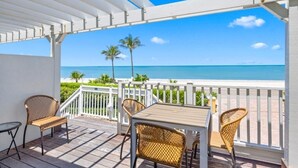 A covered outdoor area with a table and chairs, overlooking a beach with palm trees and the ocean. - Ark 3 Bedroom Condo by RedAwning (Captiva)