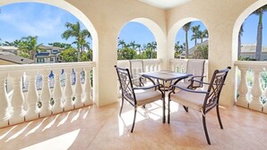 A covered outdoor patio with a table and chairs, surrounded by white balustrades and arches, overlooking a residential area with palm trees.