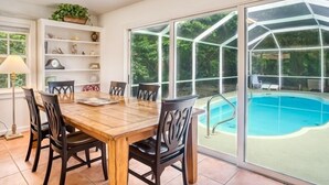 A dining area with a wooden table and chairs, a bookshelf, and a large window overlooking a swimming pool.
