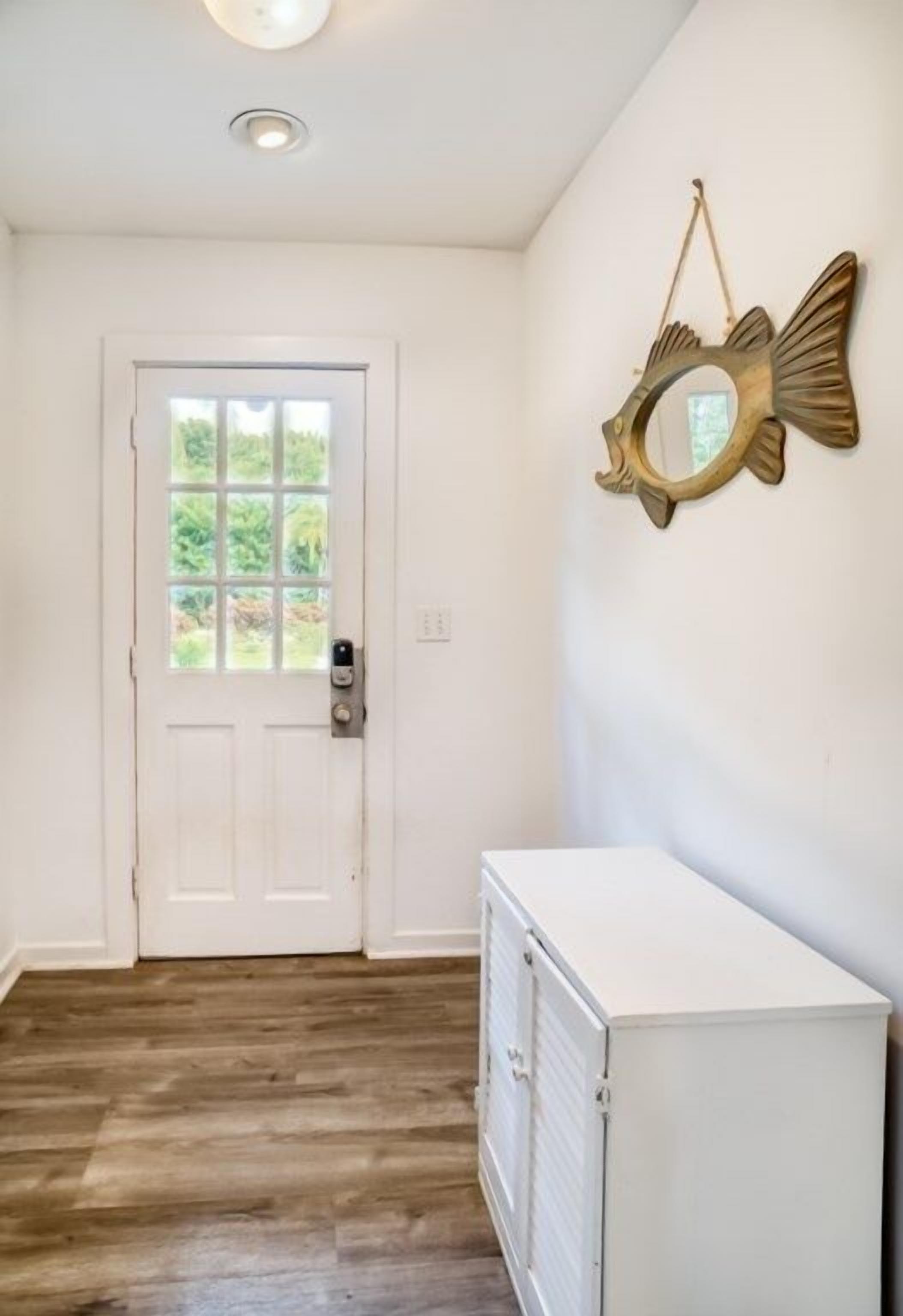 Un couloir avec une porte blanche, un miroir en forme de poisson accroché au mur et un meuble blanc.