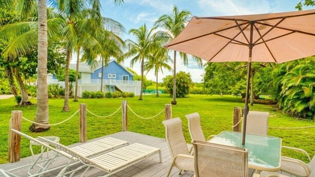 A wooden deck with a table and chairs, surrounded by a rope barrier, palm trees, and a blue house in the background.