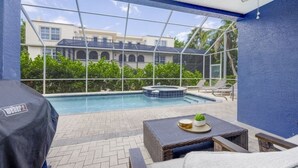 Een zwembadterras met een overdekte barbecue, ligbedden en uitzicht op een gebouw met zwembad en jacuzzi.